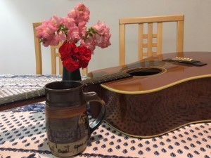 flowers in a vase, mug, and guitar sitting on a blue and white table cloth.