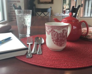 Red teapot, tea cozy, teacup, and journal and pen sitting on a wooden table. 