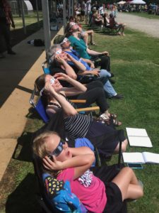 Row of people sitting in chairs holding their eclipse glasses on and gazing at the sky