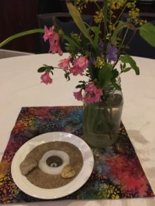 A mason jar with pink wildflowers and some greenery along with a small bowl of sand holding a tea light candle all sitting on colorful rectangle of fabric resting on a white table