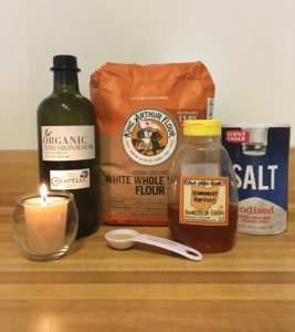 Bread baking ingredients sitting on a kitchen counter: olive oil, bag of whole wheat flour, honey, salt, measuring spoon with yeast, a beeswax vigil candle burning.