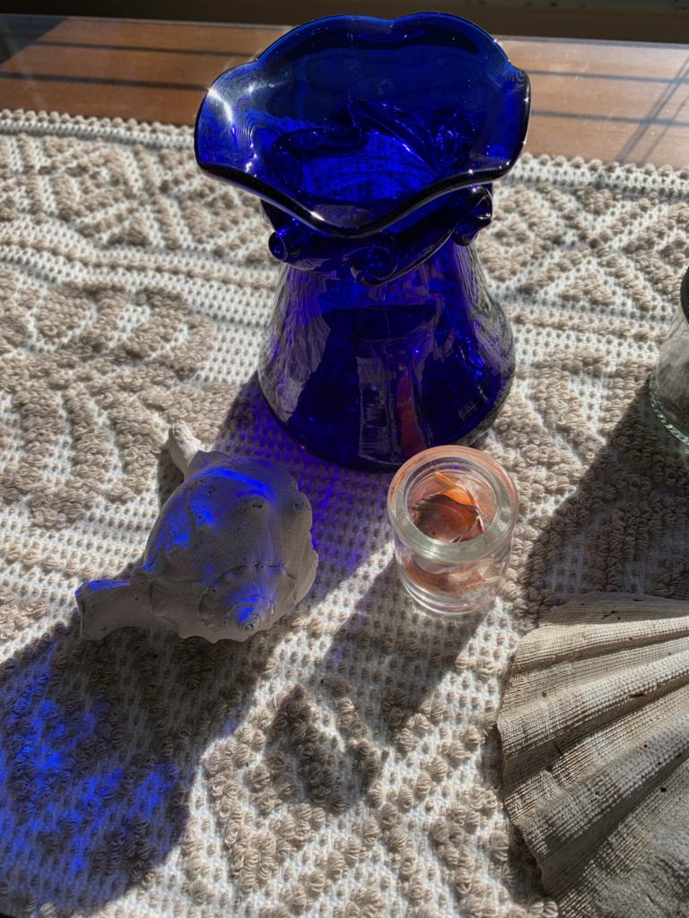 Close-up of cobalt blue vase, filtering bright sunlight and casting blue hue on sea shells and fabric runner on top of buffet